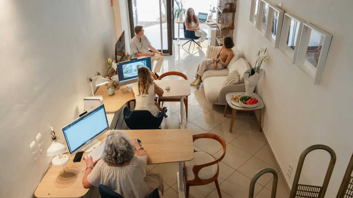 An overhead view of the studio with the whole team at work — architects at their desks and colleagues reviewing plans by the window.