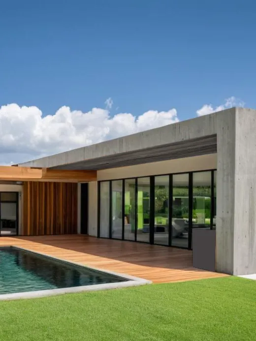 Low single-storey house in board-formed concrete and warm timber, with a dark-lined pool, lawn and mountain backdrop.
