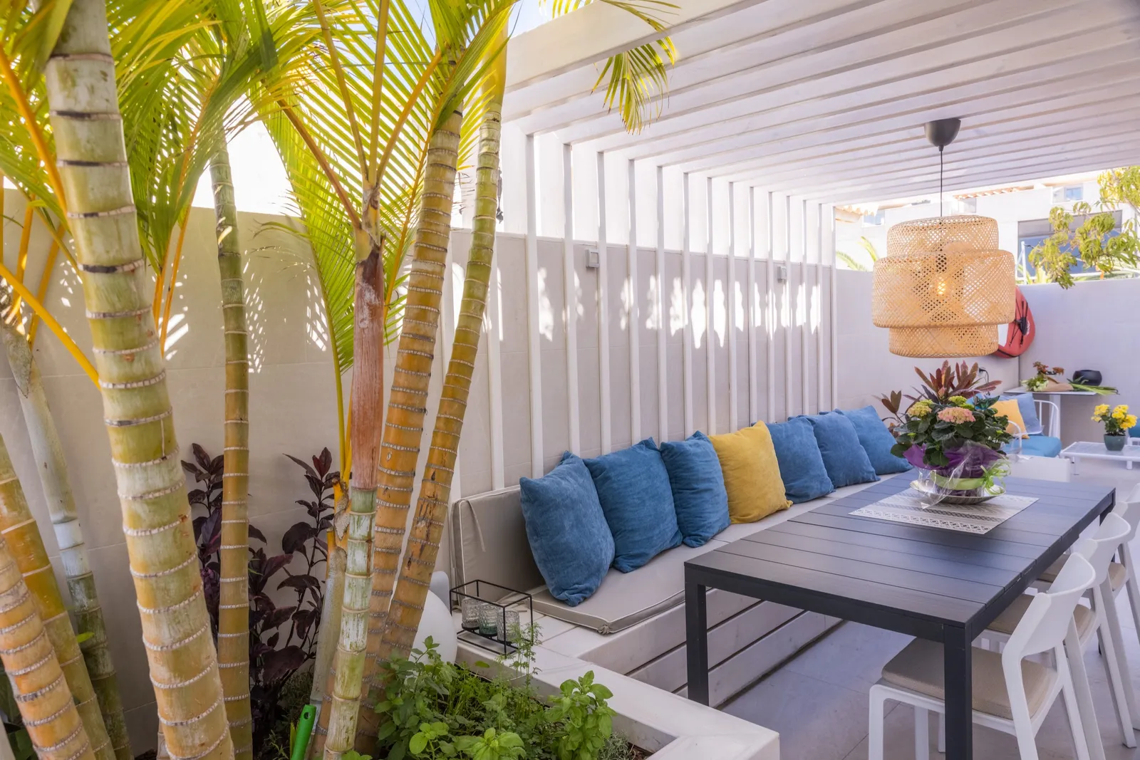 Pergola-shaded terrace with a built-in bench, blue and yellow cushions, a dark dining table and palms.