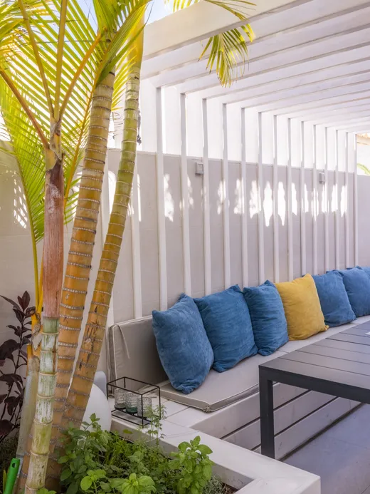 Pergola-shaded terrace with a built-in bench, blue and yellow cushions, a dark dining table and palms.