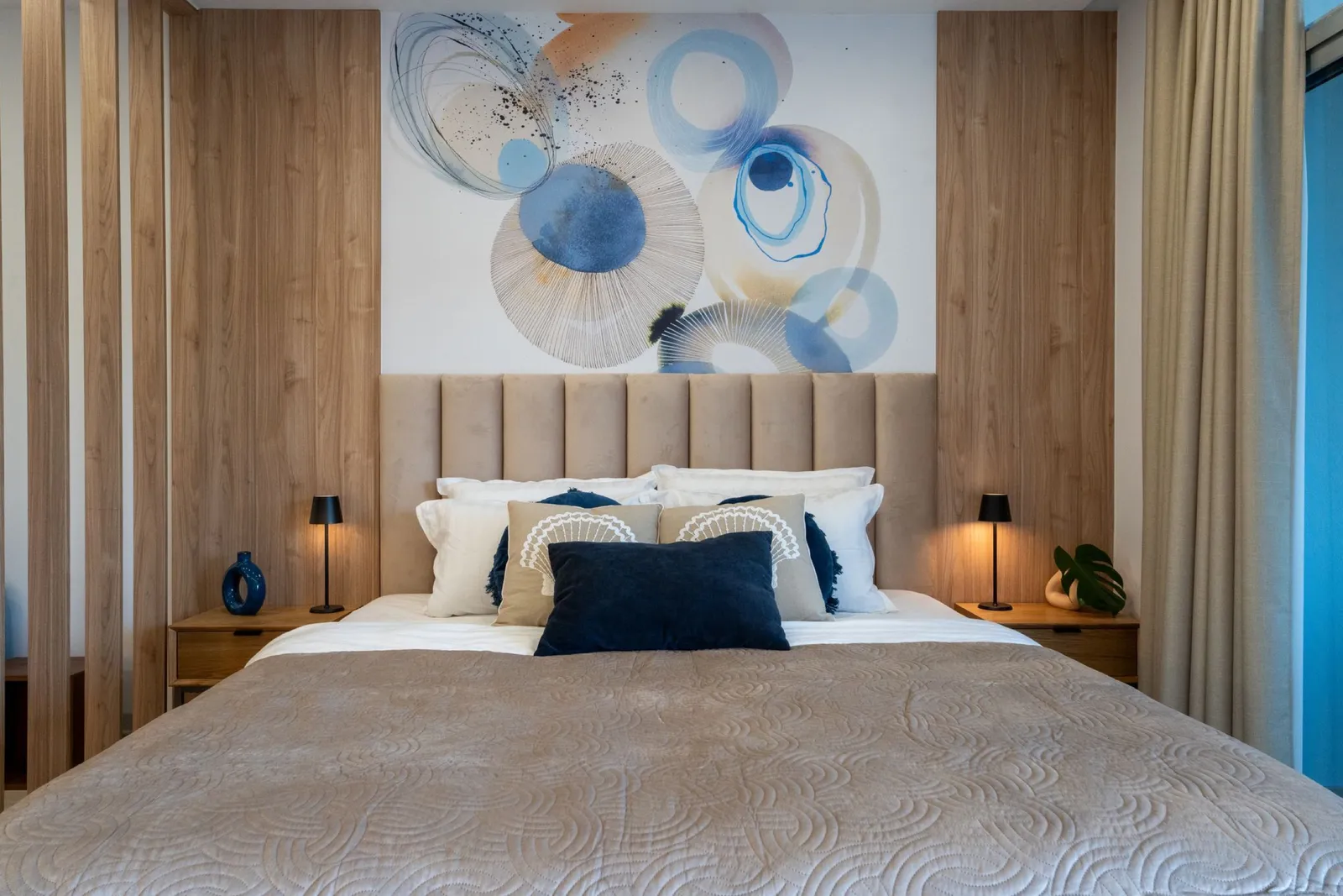 Dormitorio con un cabecero de madera y un mural abstracto circular en azul y dorado, flanqueado por lámparas negras.