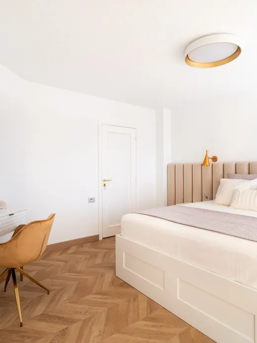 Bedroom with an upholstered headboard, a white desk, a round mirror and herringbone floors.