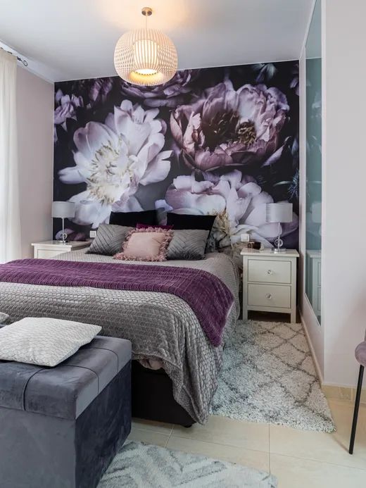 Bedroom with a dark floral peony mural, purple and lilac bedding, a velvet bench and a mauve chair.