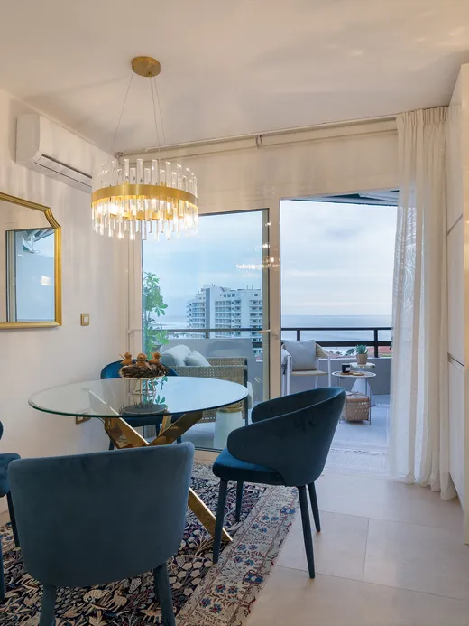 Glass dining table with teal velvet chairs, a gold mirror and a sea-view balcony beyond.