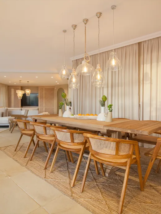 Timber dining table with woven cane chairs beneath glass pendants, the living room beyond.