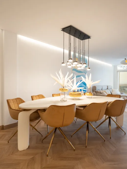 Dining area with a white oval table, tan leather chairs and a sculptural coral-branch pendant light over herringbone floors, with a sea view.