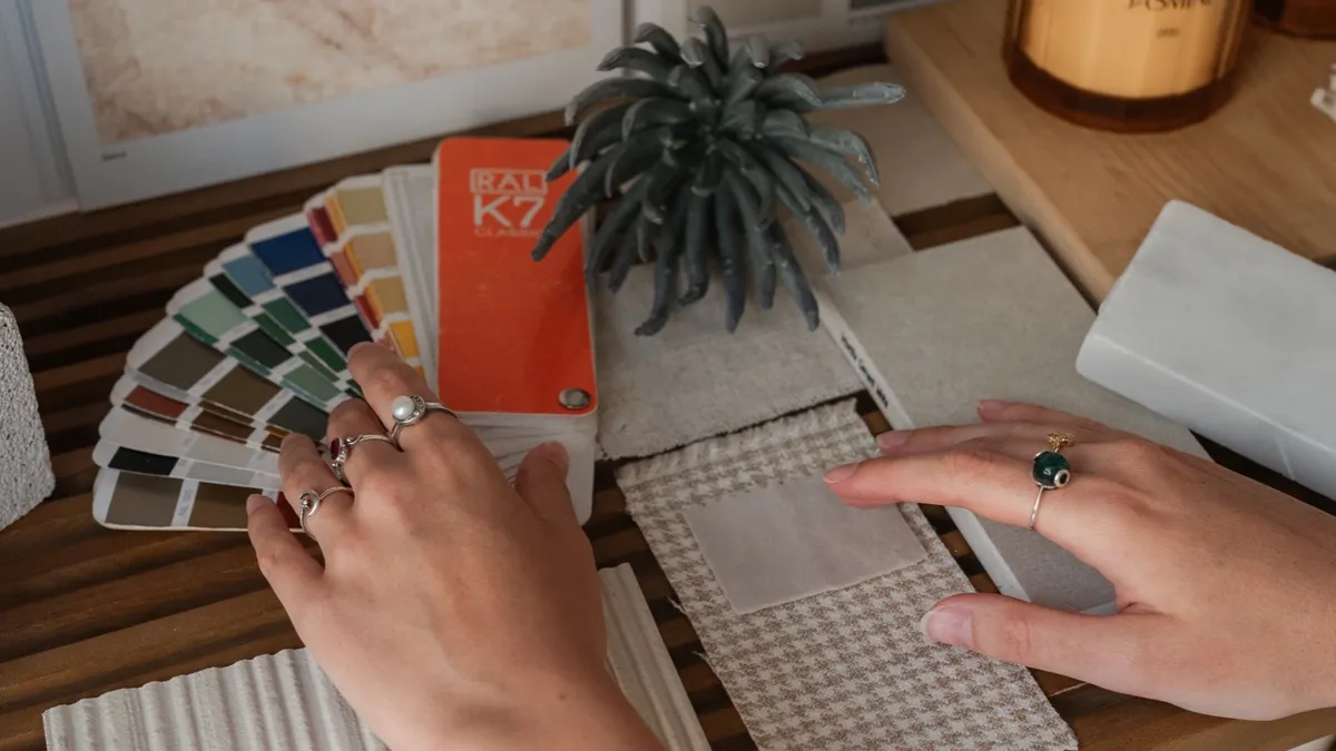 A close-up from above of hands sorting fabric swatches, a colour fan, and tile samples laid out on a desk.
