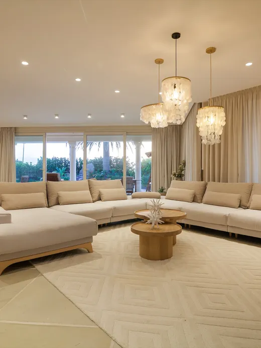 Spacious neutral living room with cream sofas, capiz-shell chandeliers and full-height drapes opening to a garden.