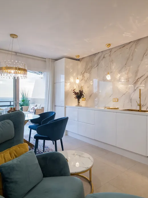 Living area with a marble feature wall, brass fixtures, teal armchairs and a sea view.