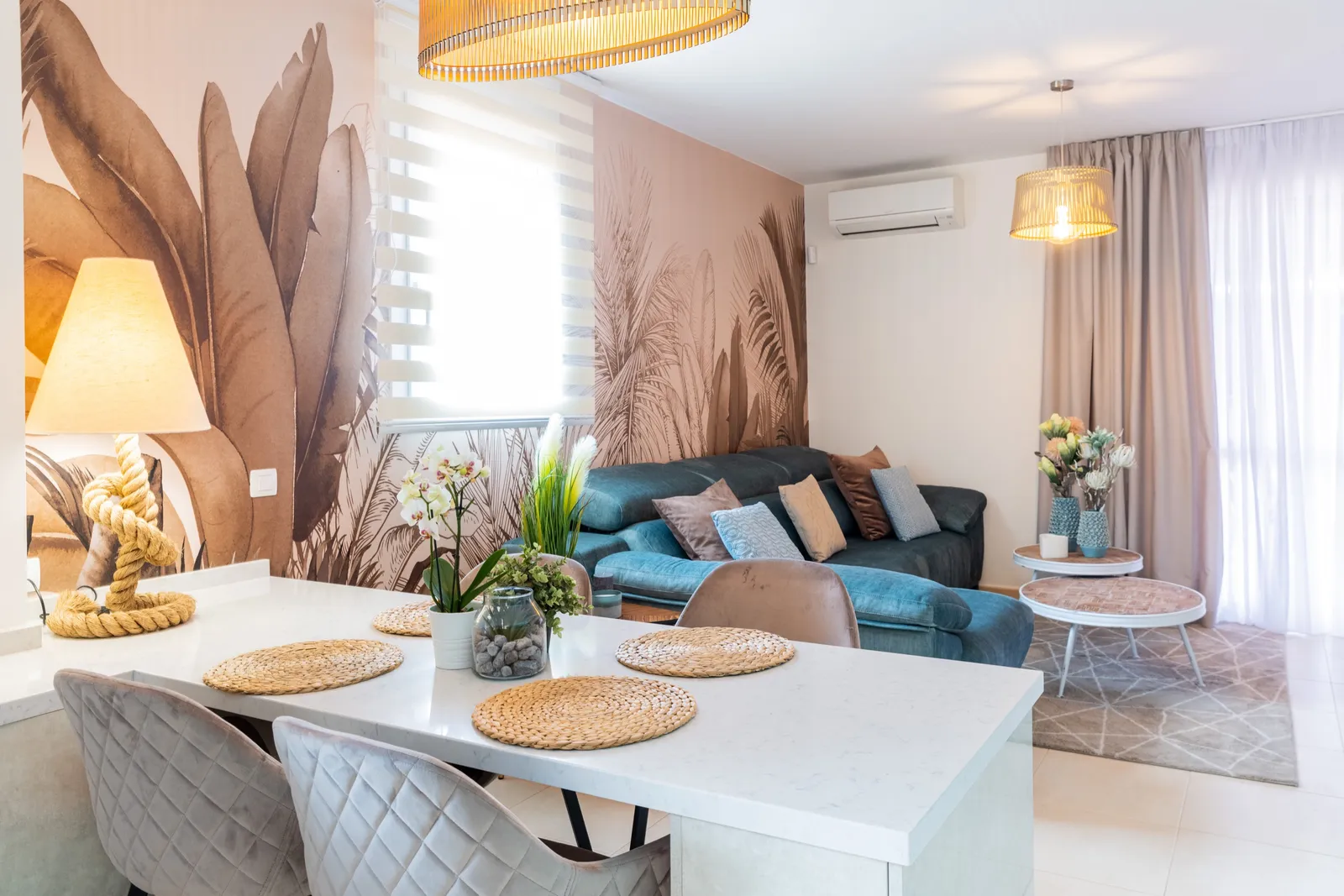 Otwarty salon z jadalnią z tropikalnym muralem palm w miękkim brązie, niebieską sofą i białą wyspą kuchenną.