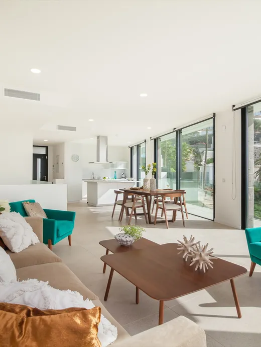 Open-plan living room with a linen sofa, turquoise accent chairs, camel leather cushions and a timber dining table, opening through full-height glazing to the garden.