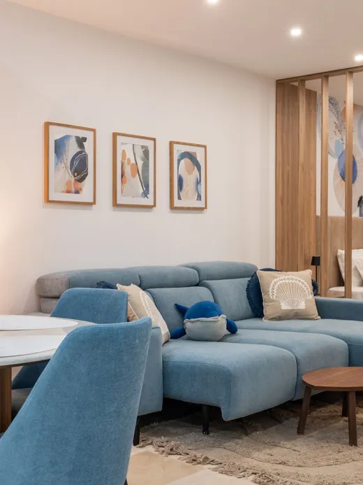 Living room in soft blues and sand tones, with a blue sofa, an oak slat divider, framed abstract art and white flowers.