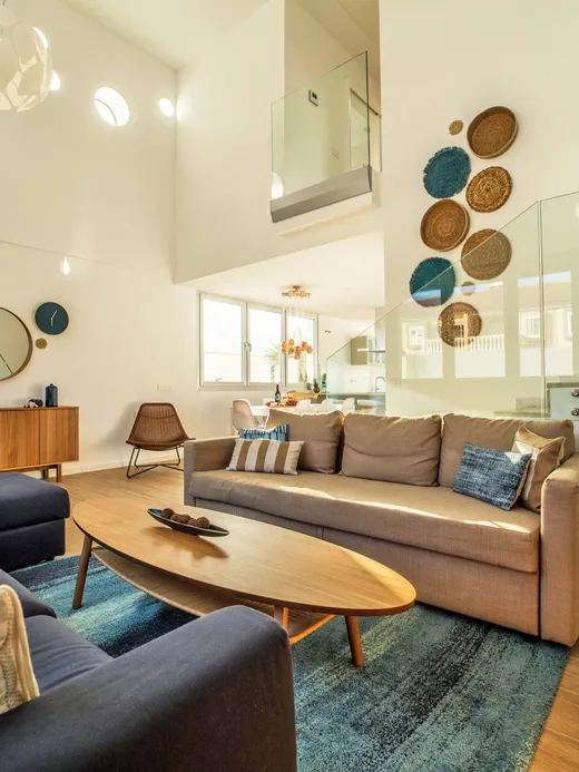 Double-height living room with a floating staircase, glass balustrade, a blue rug and woven wall discs, open to the kitchen and dining area.
