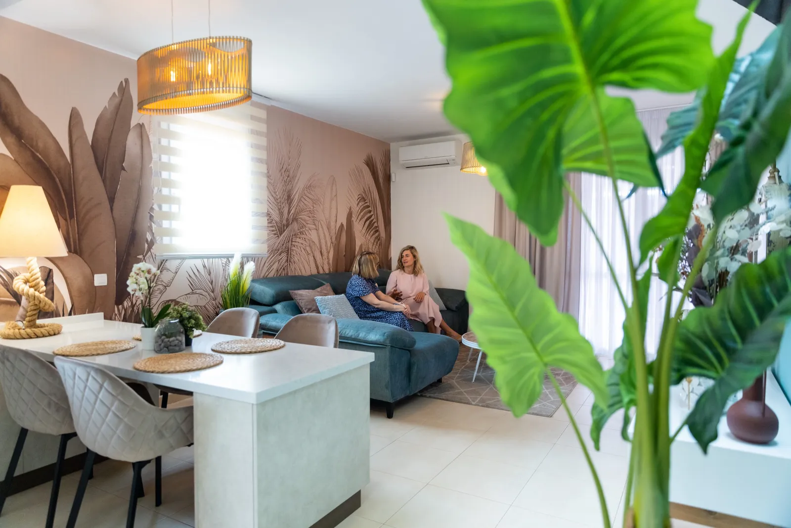 Strefa salonu i jadalni z tropikalnym muralem palm, niebieską sofą i dużą liściastą rośliną.