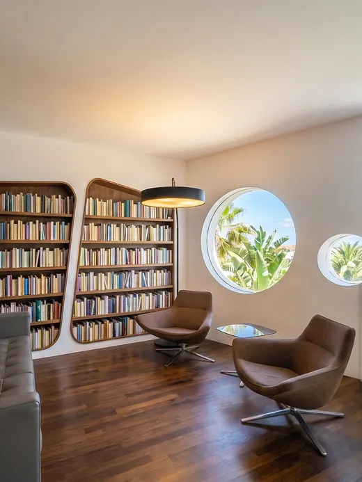Upstairs reading nook with built-in walnut bookshelves, leather sofa, and round porthole windows framing garden and sky views.