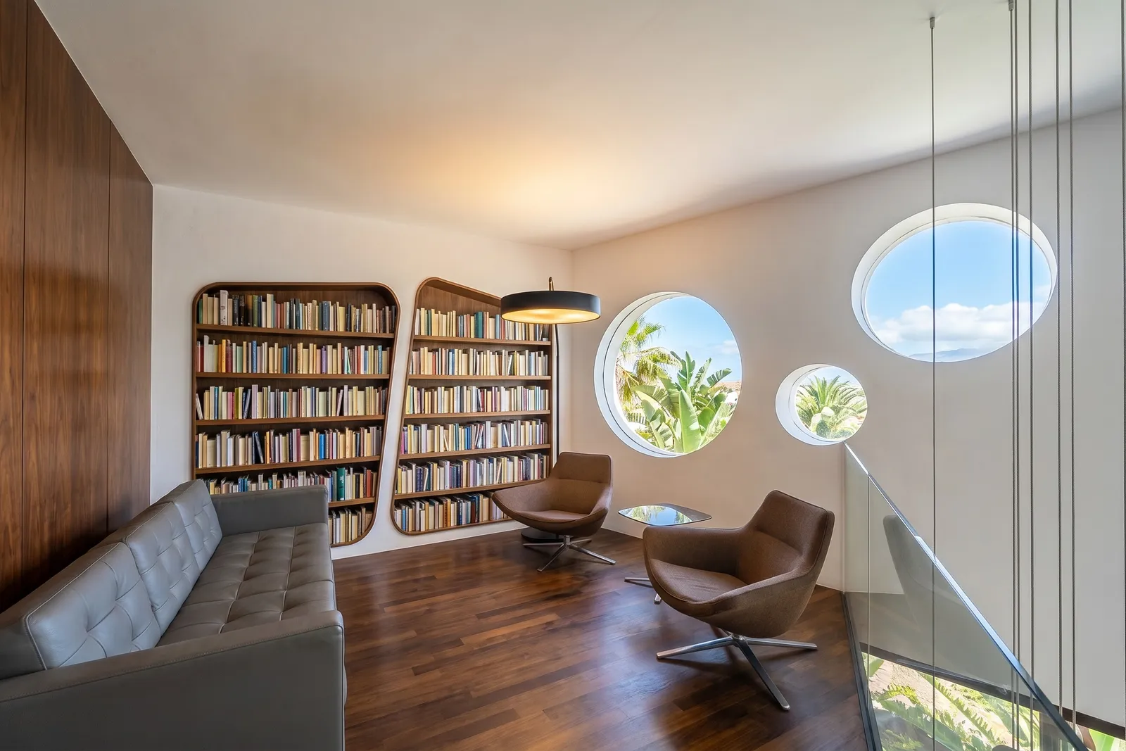 Rincón de lectura en la planta superior con estanterías de nogal empotradas, sofá de piel y ventanas circulares tipo ojo de buey con vistas al jardín y al cielo.